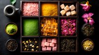 Colorful tea ingredients and additives in wooden box compartments with pink rose petals, green matcha powder, brown sugar cubes, and spices on dark background.