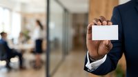 Businessman in dark suit holding blank white business card in modern office environment with blurred colleagues in background, suitable for corporate branding and professional networking.