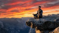 Young man meditating on mountain cliff edge during dramatic sunset with orange and pink sky colors over valley landscape for wellness and mindfulness concepts.