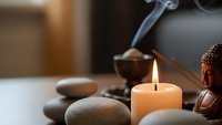 Zen spa meditation setup with burning candle, smooth stones, Buddha statue and incense smoke creating peaceful wellness atmosphere.