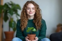 Young Caucasian woman with long curly ginger hair wearing green sweater holds potted plant, sitting cross-legged and smiling at camera. Indoor lifestyle portrait.