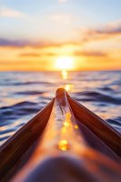 Wooden kayak bow pointing towards golden sunset over ocean waves, with sun reflection creating glowing path on water surface. Perfect for adventure travel content.