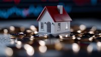 Small white model house surrounded by scattered coins on dark surface with blurred financial chart background, representing real estate investment and property market.