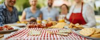 Friends gathering outdoors for casual summer meal with red checkered tablecloth, fresh food and drinks spread across table, creating warm social atmosphere.