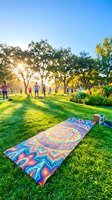 Colorful yoga mat laid on lush green grass in sunny park with trees and people exercising outdoors during golden hour, ideal for wellness and fitness marketing.