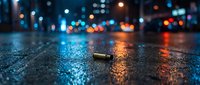 Single bullet casing lies on wet asphalt street at night with colorful city lights creating bokeh reflections on rain-soaked pavement surface.