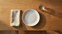 Overhead view of empty white ceramic bowl with folded linen napkin and glass of water on wooden table surface for dining and food styling.
