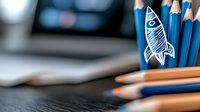 Sketch of rocket drawn on blue pencil among wooden pencils on dark desk with laptop in background, creative startup concept for business presentations.