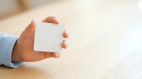 Young adult with medium skin tone holding blank white square card or paper against wooden table surface, suitable for branding, marketing mockups, and business presentations.