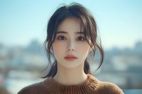 Young Asian woman with natural makeup wearing brown sweater gazes thoughtfully into distance against soft urban backdrop. Gentle daylight illuminates delicate features.