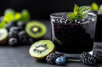 Fresh blackberry smoothie with mint garnish in glass, surrounded by kiwi slices, blueberries and blackberries on dark slate surface.