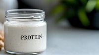 Protein powder in glass jar on kitchen counter with green plants in background for healthy nutrition and fitness lifestyle supplements.