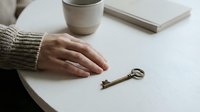 Hand reaching for vintage brass key on white wooden table with coffee cup and book in cozy minimalist setting for real estate and home concepts.