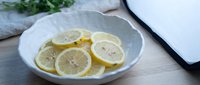 Fresh lemon slices arranged in a white ceramic bowl on a wooden surface, with green herbs and a dark tray in the background.