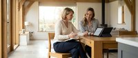 Two middle-aged professionals, Caucasian, reviewing documents and using a tablet together at a wooden desk in a bright, modern home office with large windows and natural light.