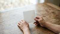 Mature man with wrinkled hands holding blank white card on wooden table indoors, suitable for greeting cards, business messaging, and promotional campaigns.