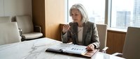 Mature woman with white hair and light complexion, wearing professional gray blazer, sitting at marble desk in modern office with city skyline view, reviewing documents and taking notes during business meeting or work session.