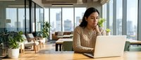 Young South Asian woman working on laptop at wooden desk in modern high-rise office with floor-to-ceiling windows, potted plants, and city skyline view. Professional setting for business, technology, and corporate themes.