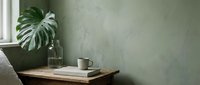 Minimalist workspace with potted monstera plant, coffee mug, and books on wooden desk against textured sage green wall near window for home office and interior design projects.