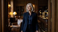 Professional mature Caucasian businesswoman with blonde bob haircut in navy blue suit standing confidently in elegant hotel lobby or upscale office interior.