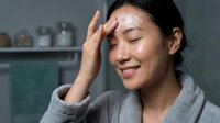 Young Asian woman applying white facial cream moisturizer on forehead in modern bathroom, skincare routine, beauty wellness self-care concept.