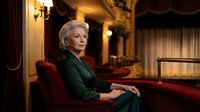 Elegant senior Caucasian woman in emerald green dress sitting in luxurious vintage interior with fireplace and ornate decor for lifestyle content.
