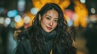 Young Asian woman with long wavy hair wearing cozy sweater against blurred city lights bokeh background at night for lifestyle and fashion content.