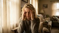 Young blonde woman experiencing headache holding head in hands sitting in bright living room with natural sunlight streaming through curtains.
