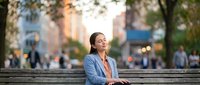 Professional businesswoman in blue blazer sitting on park bench during golden hour, enjoying peaceful moment in urban setting with blurred city background.
