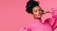 Young African American woman with elegant updo hairstyle wearing pink high neck ruffled blouse poses gracefully against pink background in fashion portrait.