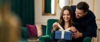 Young Caucasian couple sharing intimate moment opening blue gift box together on green sofa in cozy living room during holiday celebration.