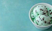 Mint chocolate chip ice cream scoops in white ceramic bowl against turquoise background, featuring creamy green base with dark chocolate pieces scattered throughout.