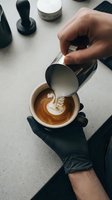 Barista pouring steamed milk into espresso cup creating swan latte art, hands in black gloves holding ceramic mug on marble counter.