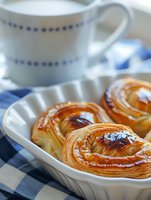 Fresh baked flaky pastries with golden crust and caramelized glaze served on white ceramic plate with coffee cup and blue checkered napkin for breakfast.