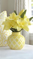 Fresh yellow daylilies arranged in geometric patterned yellow ceramic vase, complemented by matching decorative pillows and white curtains in bright interior setting.