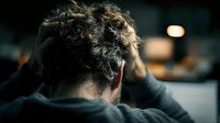 Rear view of person with curly hair holding head in hands, expressing stress or despair against dark blurred background.