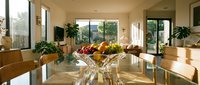 Bright modern dining room with glass table, wooden chairs, fresh fruit centerpiece, potted plants, and natural light from large windows overlooking landscape.