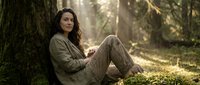 Young woman relaxing against tree trunk in peaceful forest setting with warm sunlight filtering through branches creating serene outdoor atmosphere.