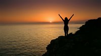 Silhouetted woman standing on rocky cliff with arms raised celebrating sunrise over ocean water at dawn, expressing freedom and joy.