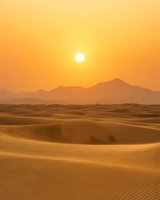 Majestic sun setting over endless sand dunes and distant mountain range creates golden hour atmosphere in vast desert landscape with rippled sandy textures.