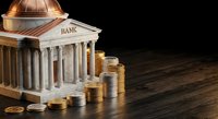 Classical bank building with golden coins stacked beside it on dark wooden surface representing financial institution wealth and monetary savings concept.