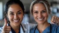 Two female healthcare workers in medical scrubs showing thumbs up and smiling at camera, representing teamwork and success in medical profession.