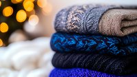 Cozy winter sweaters stack with knitted patterns in blue and gray tones against blurred warm bokeh lights background, seasonal fashion and comfort concept.