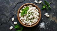 Fresh white cannellini beans in wooden bowl with mint leaves and sea salt on dark slate background for healthy cooking and Mediterranean cuisine.