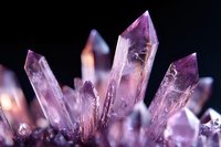 Purple amethyst crystal cluster with sharp points and natural formations against dark background, displaying ethereal glow and translucent mineral structure.