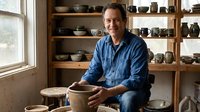 Smiling middle-aged Caucasian male potter holding a clay bowl in a rustic studio with shelves of handmade ceramic pottery in the background.