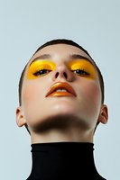 Young woman with bold yellow and orange artistic makeup looking up confidently against light blue background for fashion and beauty campaigns.