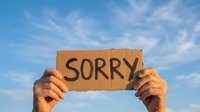 Caucasian male hands holding a cardboard sign with the word Sorry written on it against a blue sky background.