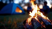 Glowing campfire with burning logs and flames in focus, blurred tent and people camping in background at night.