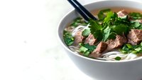 Vietnamese pho soup with beef slices, rice noodles, fresh herbs and cilantro in white bowl with chopsticks on marble surface for restaurant menu design.